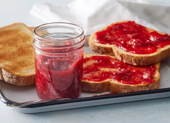 Freshly made strawberry jam in a glass jar surrounded by ripe strawberries and lemon slices on a rustic wooden table.