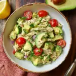 Alt text: A colorful bowl of healthy tuna avocado salad featuring chunks of tuna, creamy avocado, chopped red onions, halved cherry tomatoes, and fresh green lettuce, garnished with lemon wedges and cilantro.