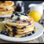 A stack of blueberry lemon ricotta pancakes topped with fresh blueberries, lemon zest, and a drizzle of maple syrup, illustrating the blueberry lemon ricotta pancakes recipe.