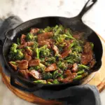 Alt text: "A savory beef and broccoli skillet recipe cooked in a cast iron skillet, featuring tender slices of beef, vibrant broccoli florets, and a rich sauce, garnished with sesame seeds and green onions."